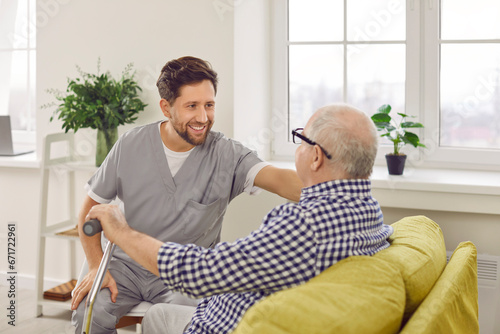 Optimistic male caregiver comforting senior patient, touching his shoulder. Male carer in uniform giving support to elderly man who sitting on sofa with walking cane. Elderly healthcare, medical care
