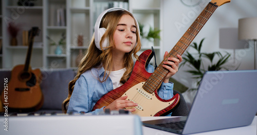 Beautiful teenager girl in earphones learning play guitar at home using online lessons on laptop. Beautiful girl with long hair play e guitar at home for online audience on laptop