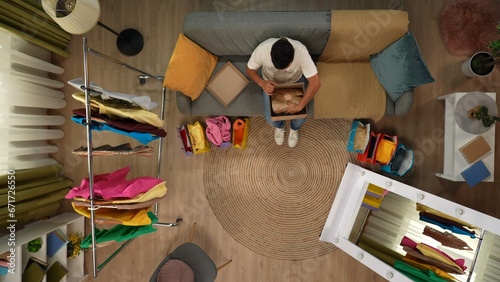 Top view of modern apartment living room. Man sitting on the sofa opens cardboard box and looks at the package he received, positive expression.