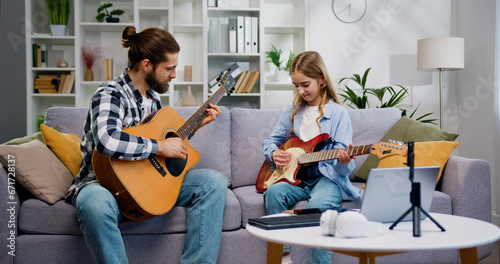 Young musician teacher and 10 year old girl sitting on the sofa in living room, playing the guitar and enjoying music while having music class together, indoors