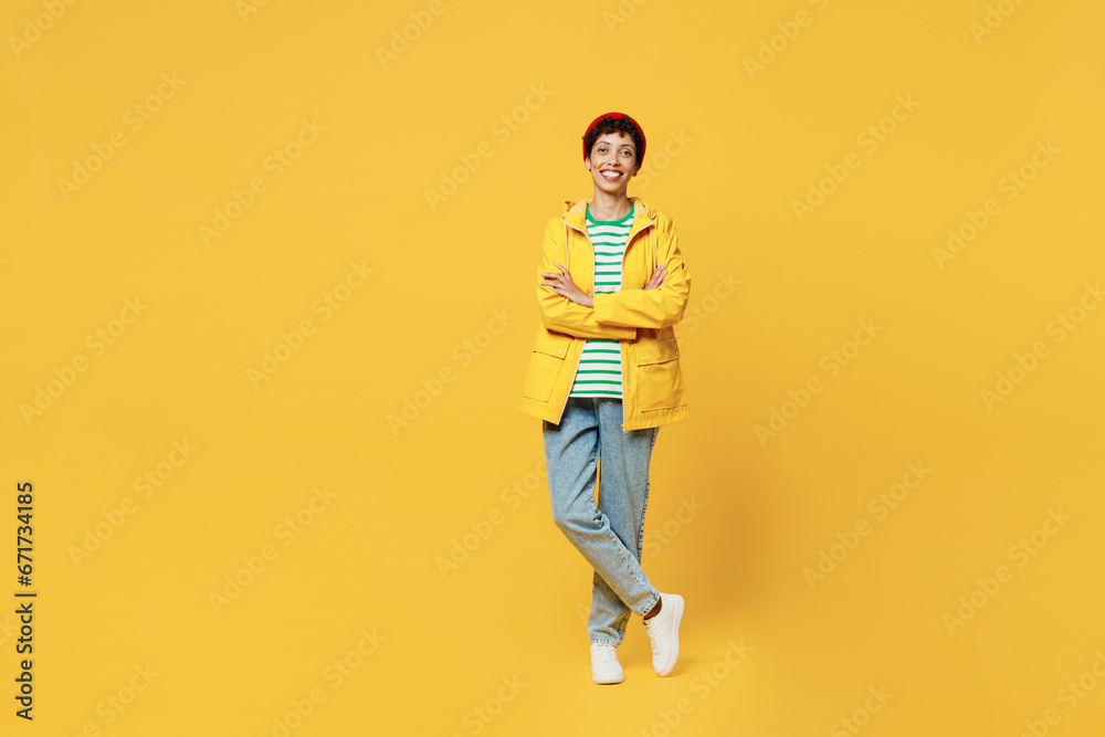 Full body young fun woman wears waterproof raincoat outerwear red hat hold hands crossed folded isolated on plain yellow background studio portrait. Outdoors lifestyle wet fall weather season concept.