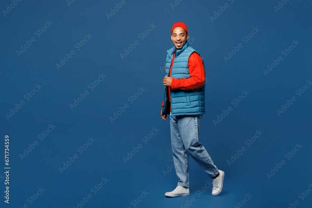 Naklejka premium Full body side view fun happy young man of African American ethnicity wear padded vest red hat walk go look camera isolated on plain dark royal navy blue background studio portrait. Lifestyle concept.
