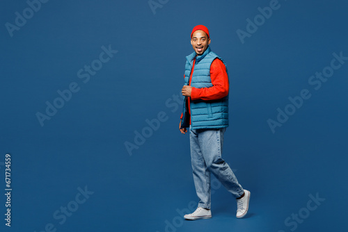 Full body side view fun happy young man of African American ethnicity wear padded vest red hat walk go look camera isolated on plain dark royal navy blue background studio portrait. Lifestyle concept.