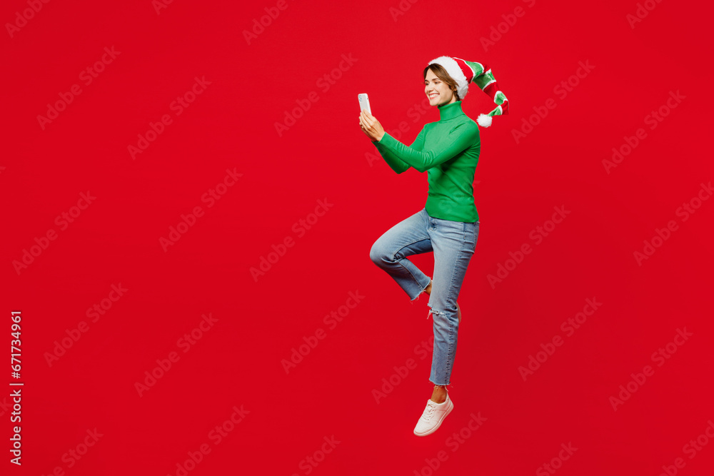 Full body young woman wear green turtleneck Santa hat posing jump high hold in hand use mobile cell phone isolated on plain red background. Happy New Year 2024 celebration Christmas holiday concept.