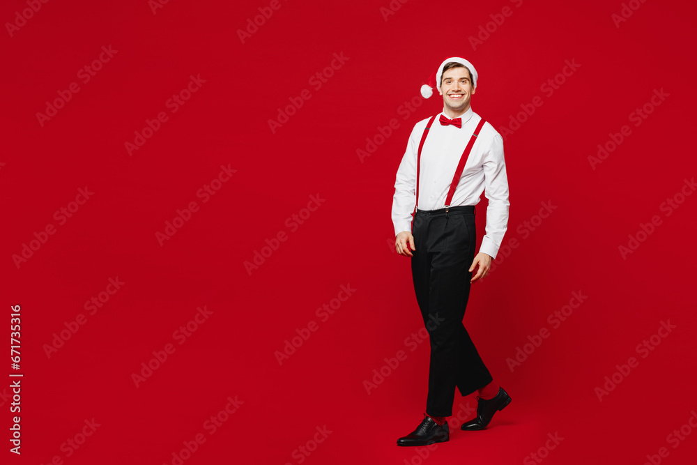 Full body side profile view merry young smiling man wear white shirt Santa hat walk go look camera posing isolated on plain red background studio. Happy New Year Christmas celebration holiday concept. Full body side profile view merry young smiling man wear white shirt Santa hat walk go look camera posing isolated on plain red background studio. Happy New Year Christmas celebration holiday concept.