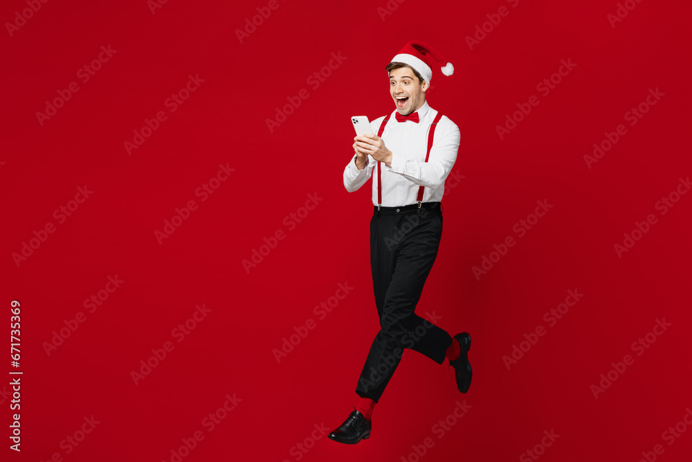 Full body sideways merry young man wears white shirt Santa hat posing jump high use mobile cell phone run fast isolated on plain red background. Happy New Year Christmas celebration holiday concept. Full body sideways merry young man wears white shirt Santa hat posing jump high use mobile cell phone run fast isolated on plain red background. Happy New Year Christmas celebration holiday concept.