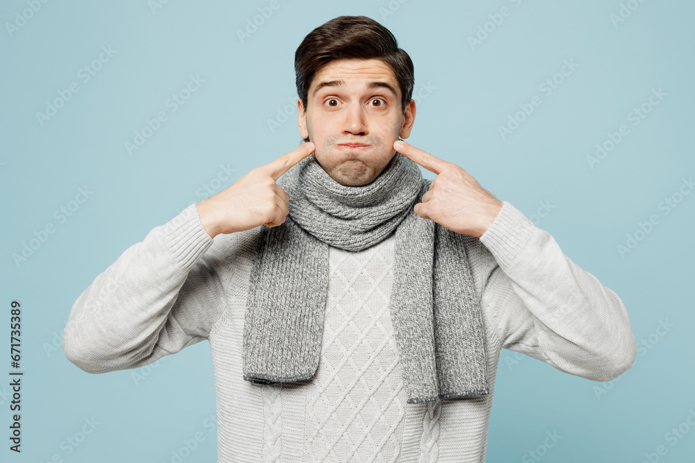 Young sick man in gray sweater scarf point fingers on blowing puffing cheeks isolated on plain blue background studio portrait. Healthy lifestyle disease virus treatment cold season recovery concept. - obrazy, fototapety, plakaty 