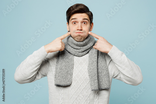 Young sick man in gray sweater scarf point fingers on blowing puffing cheeks isolated on plain blue background studio portrait. Healthy lifestyle disease virus treatment cold season recovery concept. photo