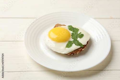 Plate with tasty fried egg and slice of bread on white wooden table