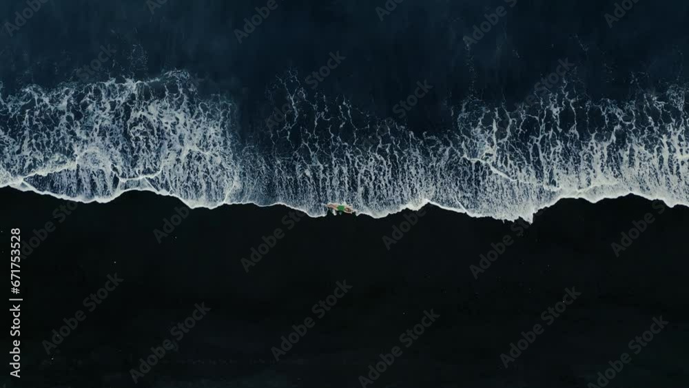 Aerial top view of a girl in a swimsuit lying on a black lava beach on the surf line with white waves. Coast of the island of Bali, Black sand beach, Indonesia.