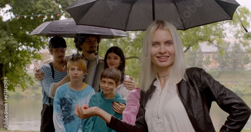 A group of joys pose under an umbrella in rainy weather. Blonde woman touches an umbrella, smiles. Positive mood in a friendly company.