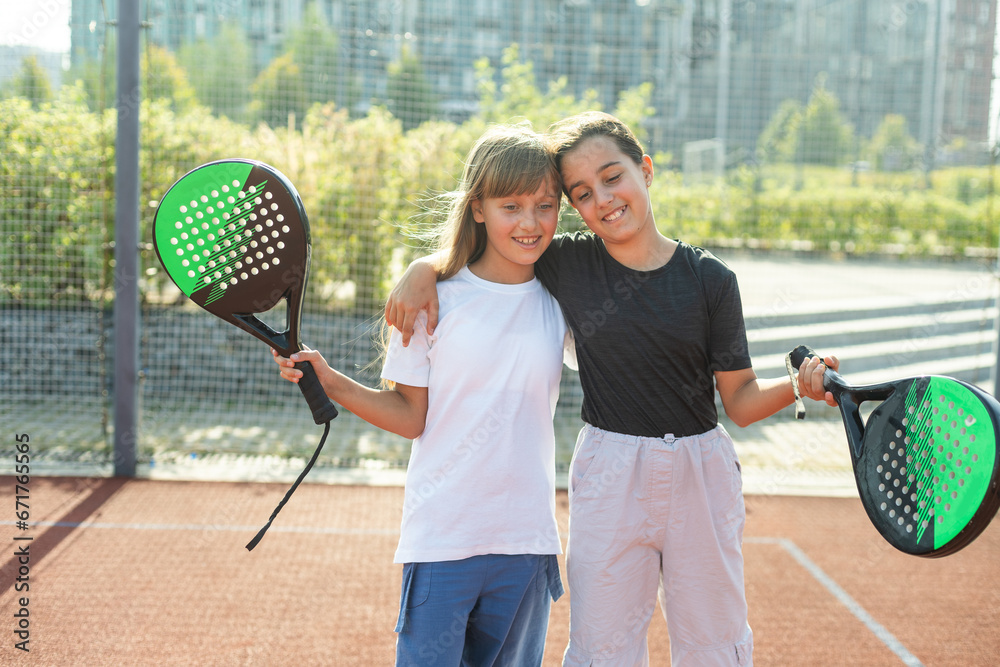 Teenage girls with racquets and balls standing in padel court, looking at camera and smiling.