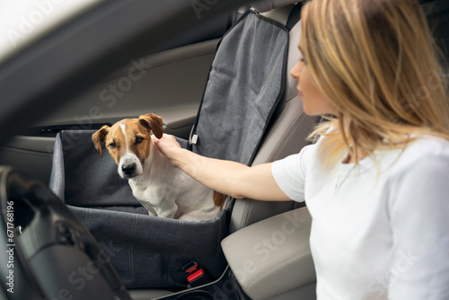 Safe Road trip by car. Petting cute dog with serious face. Dog sitting in car chair hammock on front seat of the car. Blonde young driver girl petting her pet. Focus on dog, girl is blurred