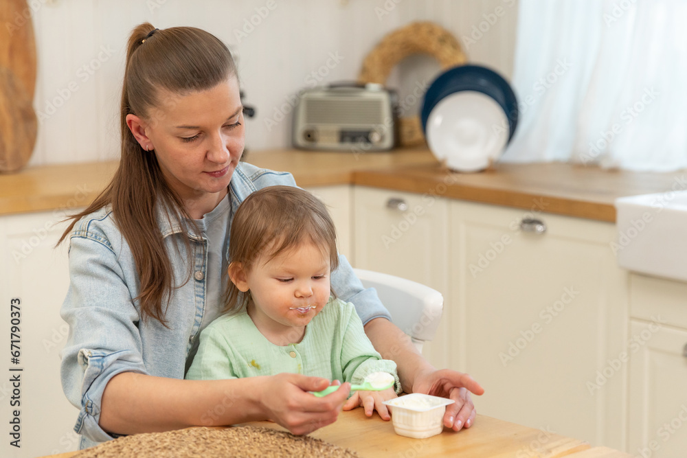 Happy family at home. Mother feeding her baby girl from spoon in kitchen. Little toddler child with messy funny face eats healthy food at home. Young woman mom giving food to kid daughter Happy family at home. Mother feeding her baby girl from spoon in kitchen. Little toddler child with messy funny face eats healthy food at home. Young woman mom giving food to kid daughter