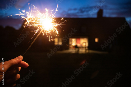 Hand holding a sparkler, or bengal fire stick, burning in outdoor setting at blue hour dusk with a house in the background. Holidays, magic wallpaper.