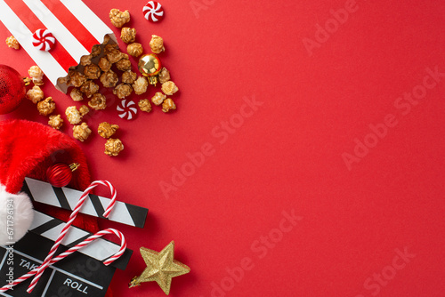 Christmas-themed movie premiere idea. Overhead shot of film clapper, delectable popcorn in striped box, Santa's hat, festive ornaments, star decor, sweet treats on red backdrop with space for text