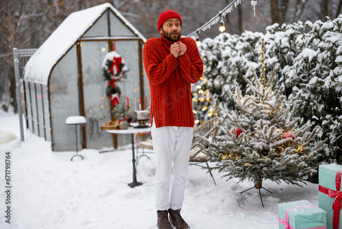 Man in red sweater and hat warms his hands while feeling cold at snowy backyard decoarted for holidays. Concept of winter time and cold weather photo