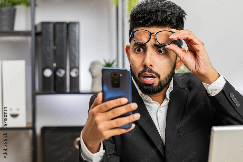 Indian businessman freelancer use smartphone typing browsing, loses becoming surprised sudden lottery results, bad news, fortune loss, fail working at home office workplace desk. Male Hindu in suit Indian businessman freelancer use smartphone typing browsing, loses becoming surprised sudden lottery results, bad news, fortune loss, fail working at home office workplace desk. Male Hindu in suit