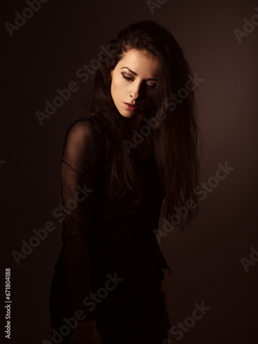 Beautiful woman looking down with brown long hair on dark shadow background. Closeup portrait.