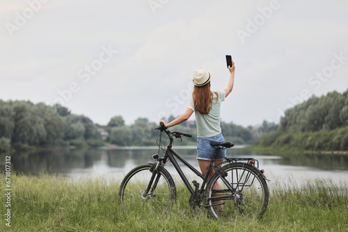 Fototapeta Naklejka Na Ścianę i Meble -  A young woman in casual clothes takes a selfie with a bicycle in a park near the river.