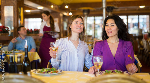 Friendly meeting of two women over dinner with wine in restaurant. Adult European and Asian female friends enjoying evening meal and carefree conversation
