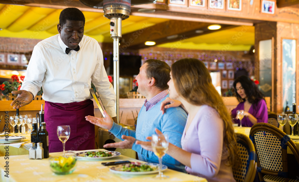 Dissatisfied guests conflicting with bewildered african american waiter because of poor quality of dishes in restaurant..