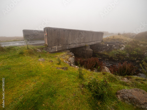 Small simple concrete bridge for one car over a river or creek. Fog in the background. Driving in dangerous conditions due to poor visibility. Connemara, Ireland. Rural country side area.