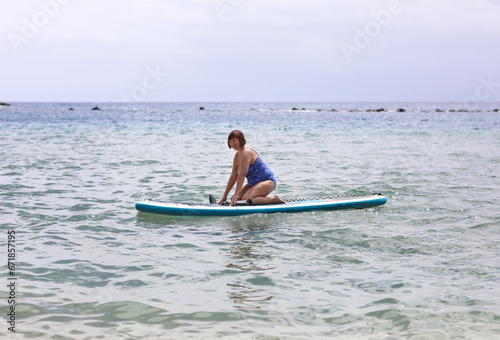 Middle aged woman in swimsuit sits on inflatable SUP board and paddling at calm ocean water. Active female enjoys does yoga, stretching or trains on paddle board at summer vacation. Healthy lifestyle