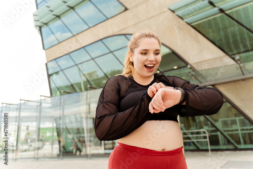 Smiling young woman checking heart rate with smartwatch while training in the city. Sport, plus size, motivation