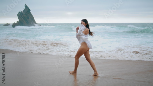 Girl stepping wet sand ocean beach wearing sexy white swimsuit cloudy day.