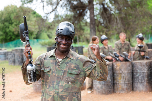 Smiling afro american man paintball player in camouflage posing with gun outdoors