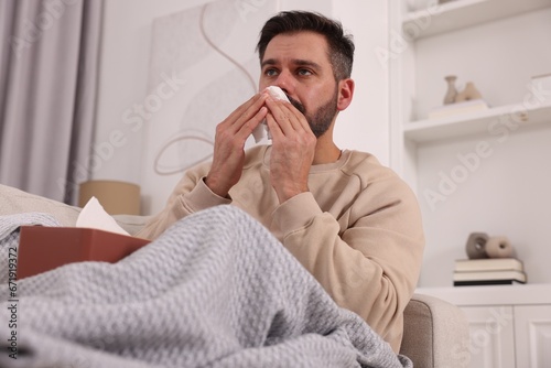 Sick man with tissue blowing nose on sofa at home. Cold symptoms