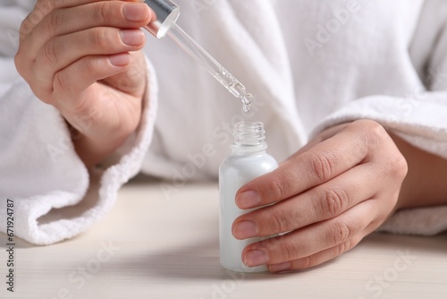 Woman dripping cosmetic serum from pipette into bottle at white table  closeup