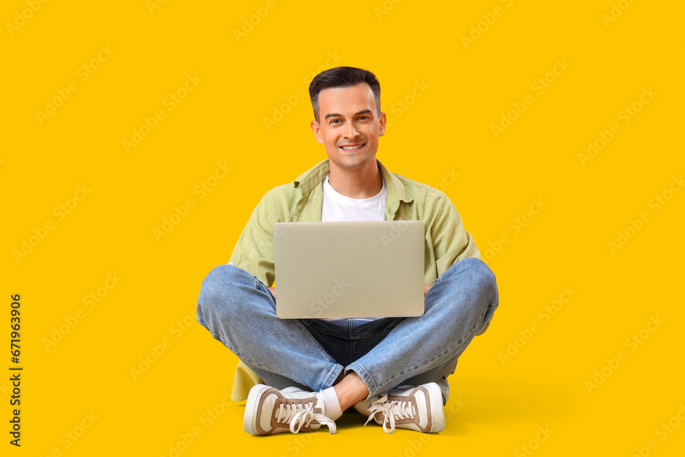 Happy young man sitting on floor with laptop against yellow background