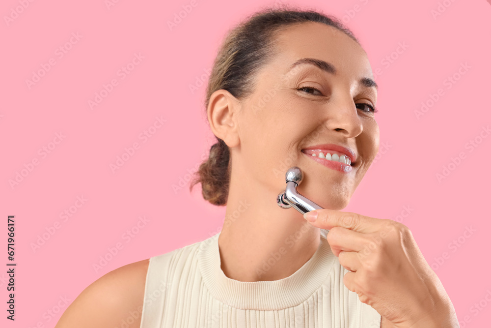 Beautiful mature woman using face massage roller on pink background Beautiful mature woman using face massage roller on pink background