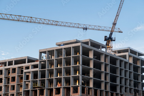 A crane and a building under construction against a blue sky background. Builders work on large construction sites, and there are many cranes working in the field of new construction.