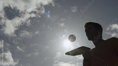 Silhouette of man using racket to hit pickleball ball against sky. Practicing curveball and spinning ball with round holes in air during training outdoors in sunny weather.