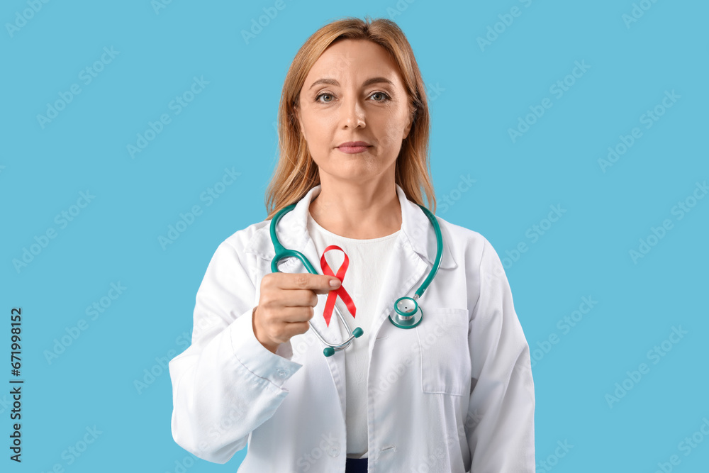 Mature female doctor with red ribbon on blue background. World AIDS Day concept Mature female doctor with red ribbon on blue background. World AIDS Day concept