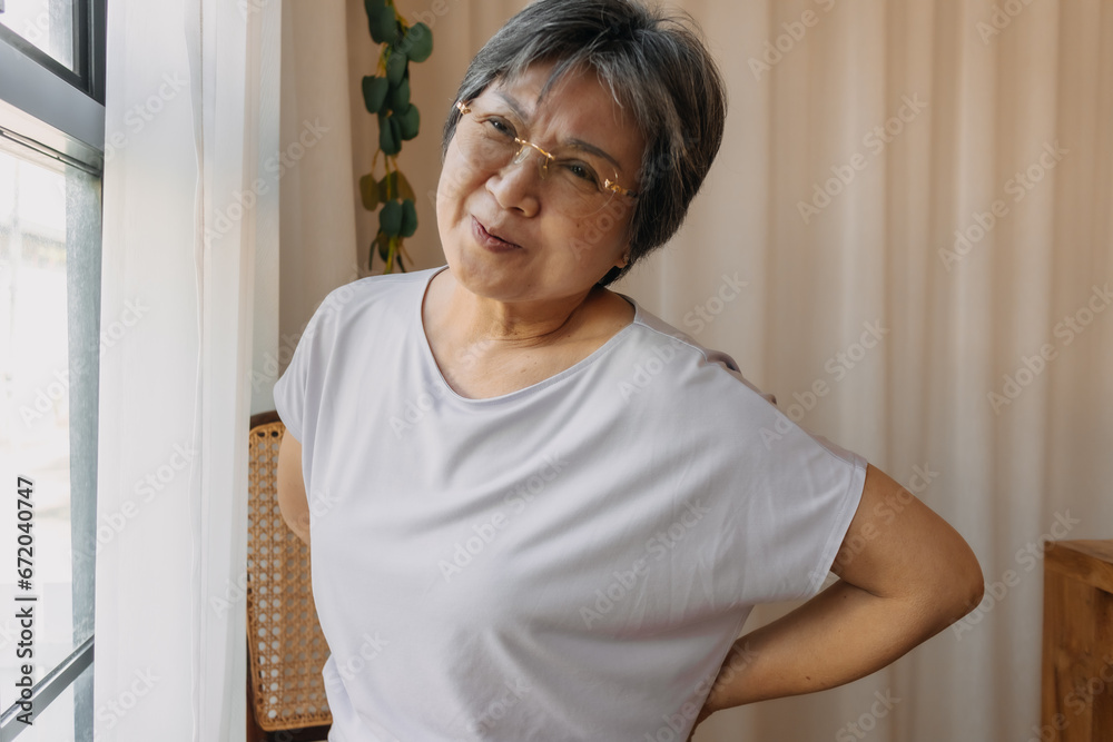 Asian old woman holding and touching lower back pain, got backache while sitting on chair next to window with painful face, groaning in pain with bad symptom. Asian old woman holding and touching lower back pain, got backache while sitting on chair next to window with painful face, groaning in pain with bad symptom.