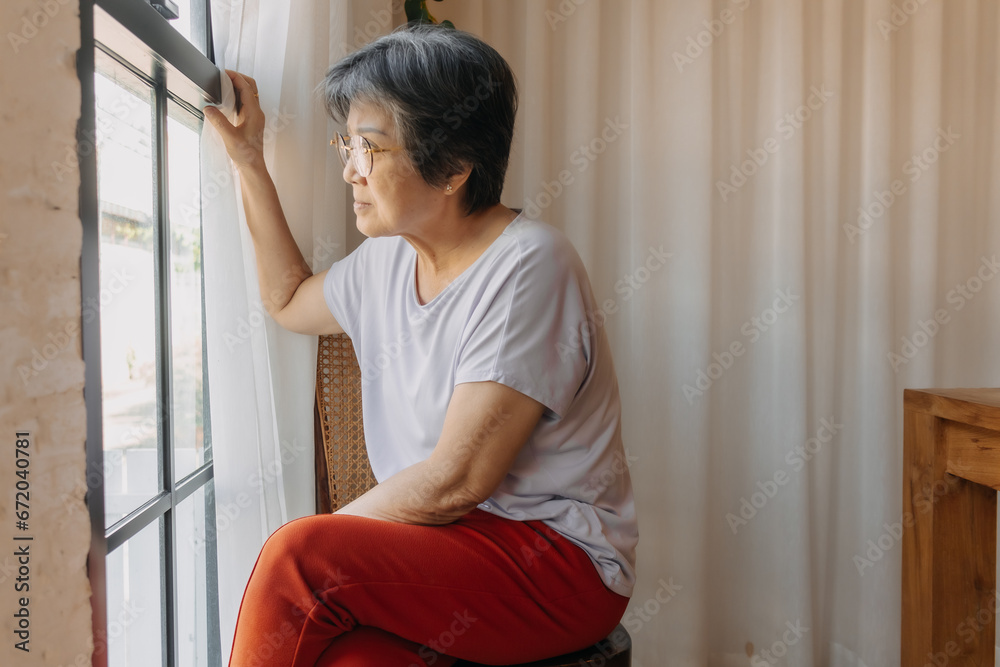 Asian old woman sitting on chair and opening white curtain while looking out the window, missing and waiting family and thinking something sad, living alone. Asian old woman sitting on chair and opening white curtain while looking out the window, missing and waiting family and thinking something sad, living alone.