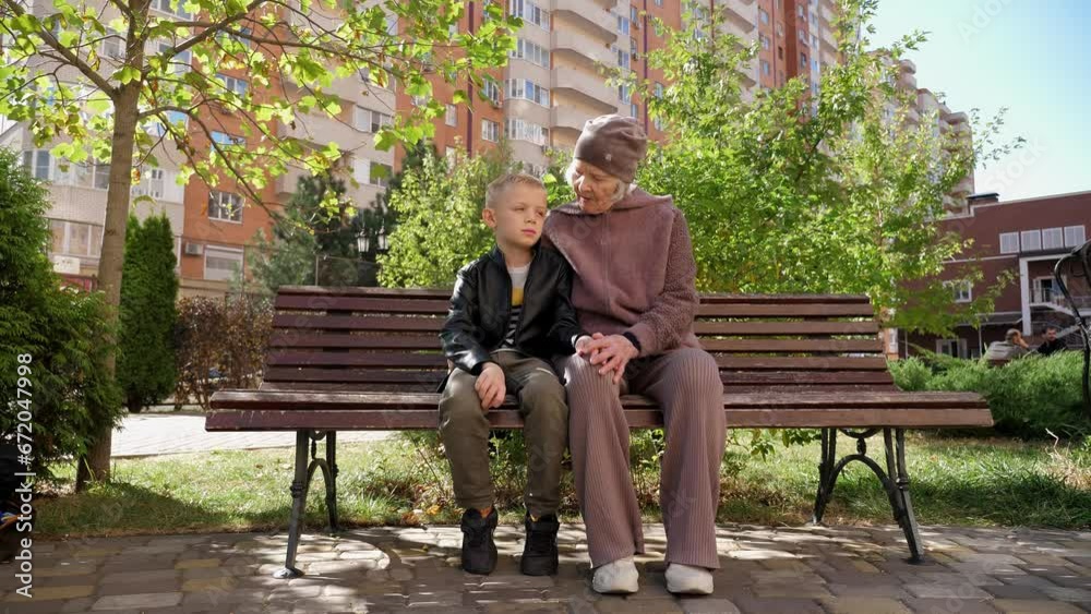 A happy caring great-grandmother spends time with her grandson sitting on a bench in sunny autumn weather, they are sitting against the background of a multi-storey modern building. A happy caring great-grandmother spends time with her grandson sitting on a bench in sunny autumn weather, they are sitting against the background of a multi-storey modern building.