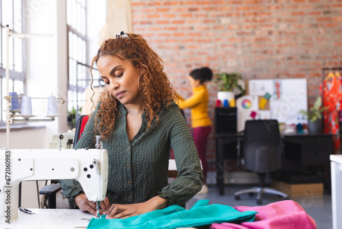 Focused biracial female fashion designer using sewing machine in sunny studio photo