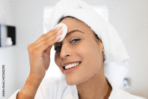 Portrait of happy biracial woman wearing white bathrobe and using cotton pad on her face in bathroom