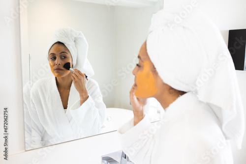 Happy biracial woman wearing bathrobe and making face mask in front of mirror in bathroom photo