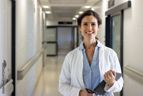 Portrait of smiling caucasian female doctor with tablet standing in hospital corridor, copy space