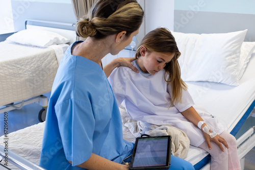 Caucasian female doctor with tablet comforting sad girl patient sitting on hospital bed photo
