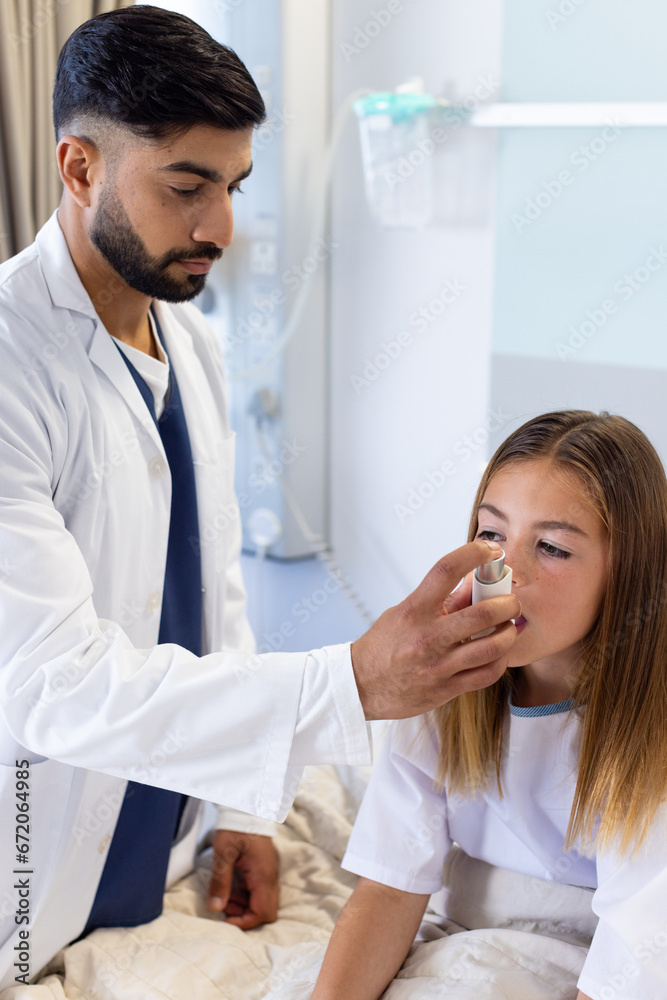 Focused diverse male doctor helping girl patient use asthma inhaler sitting up in hospital bed Focused diverse male doctor helping girl patient use asthma inhaler sitting up in hospital bed