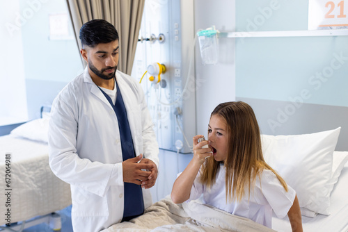 Diverse male doctor watching girl patient using asthma inhaler sitting up in hospital bed photo