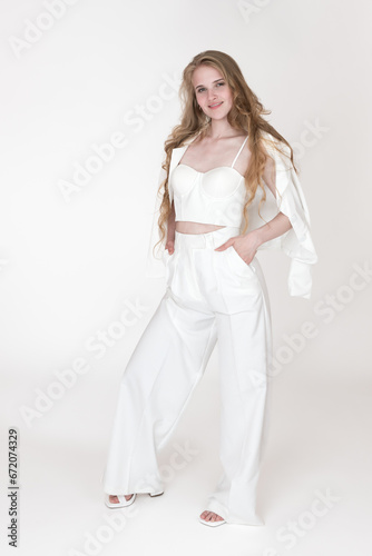 Smiling woman stylishly dressed in white suit jacket, sculpting cupped corset top, trousers, slingbacks standing on white background. Model holding hands in pants pockets, jacket draped over shoulders