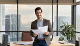 Happy young Latin business man checking financial documents in office. Smiling male professional account manager executive lawyer holding corporate tax bill papers standing at work